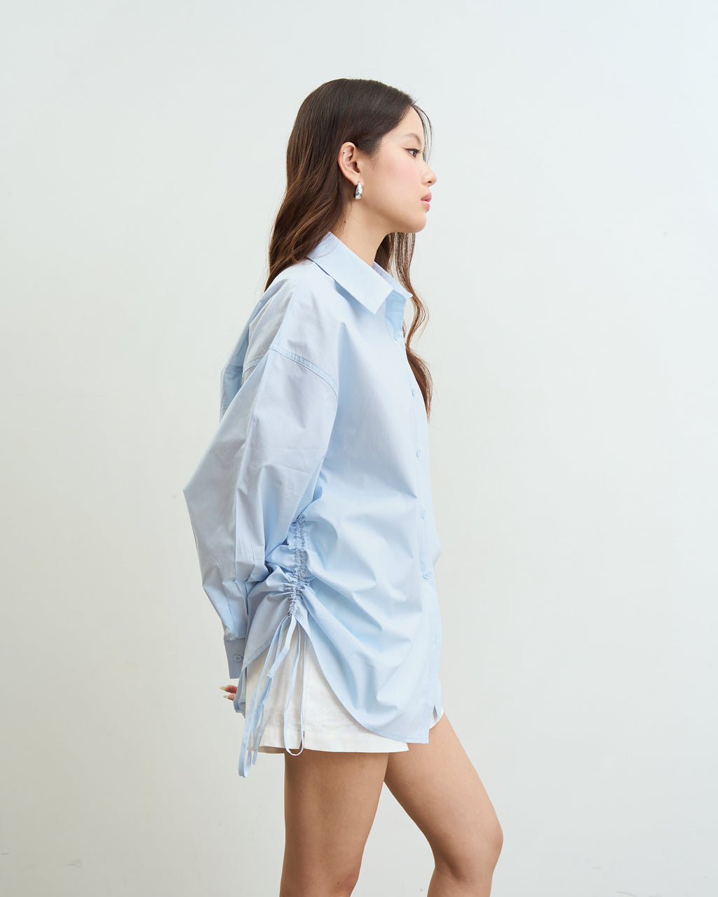 woman wearing an oversized blue button-up shirt with side drawstrings and white shorts, standing in profile against a plain off-white wall.