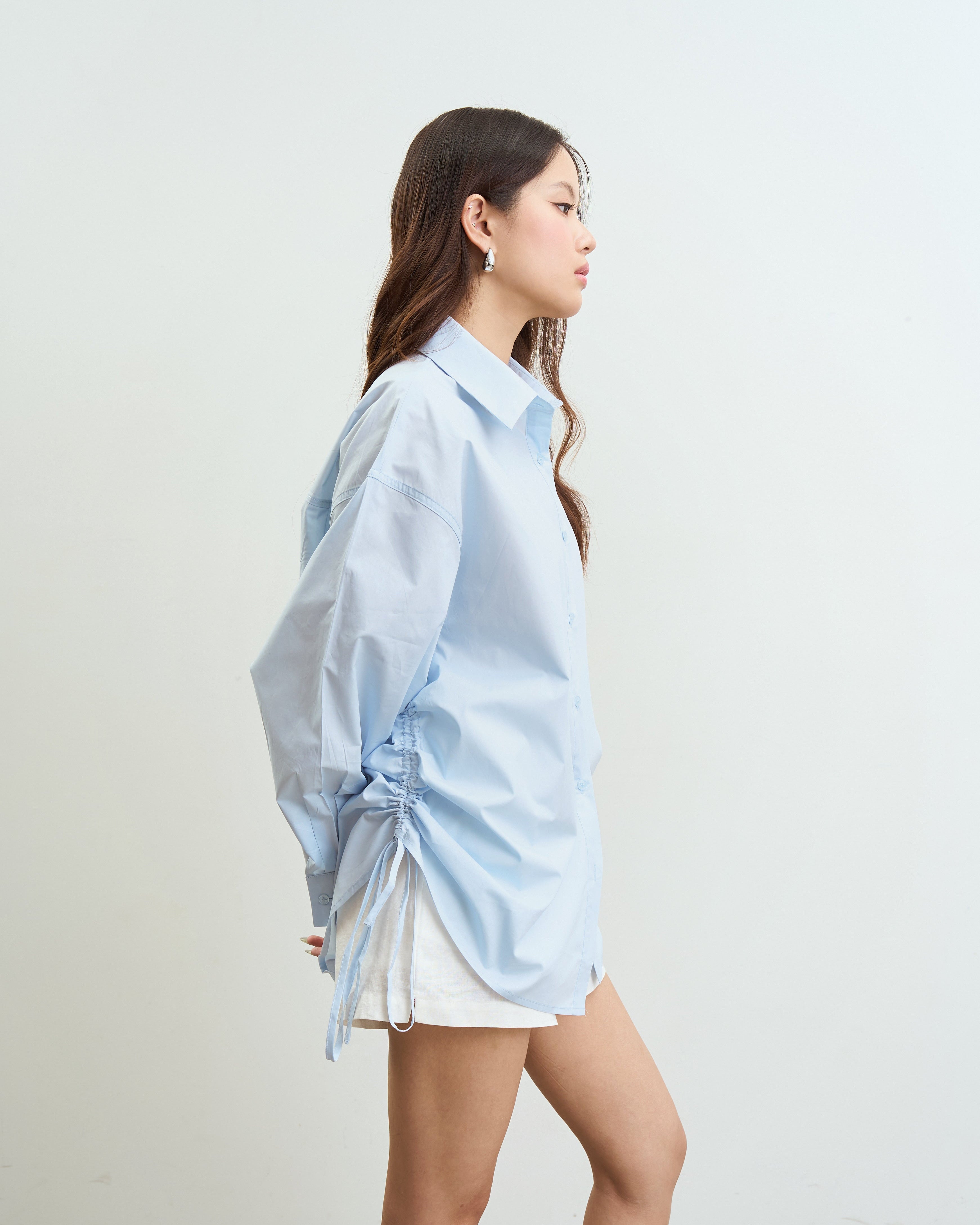 woman wearing an oversized blue button-up shirt with side drawstrings and white shorts, standing in profile against a plain off-white wall.
