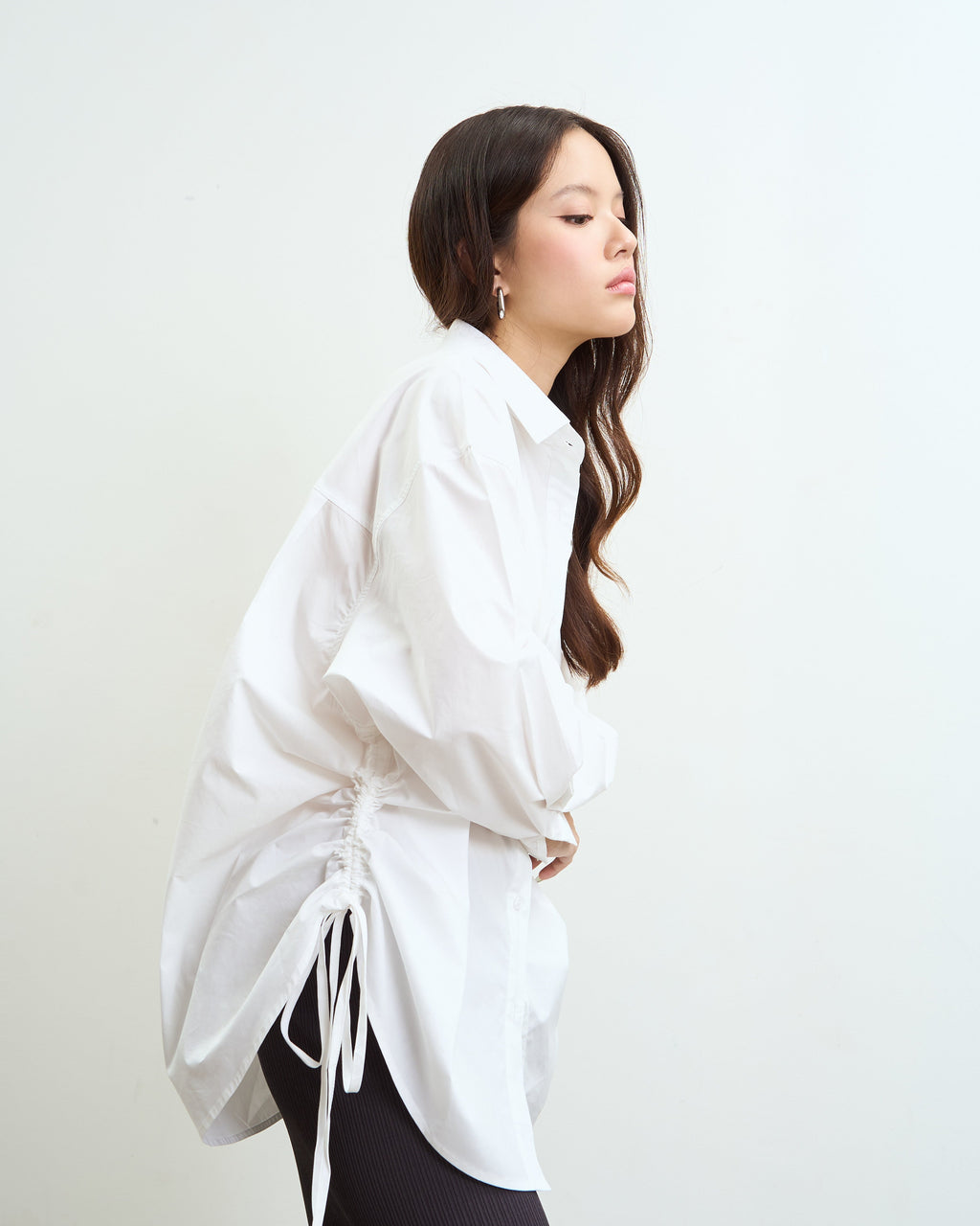 woman in profile wearing a loose white long-sleeve blouse with side drawstrings and a black ribbed skirt, photographed against a plain light background.