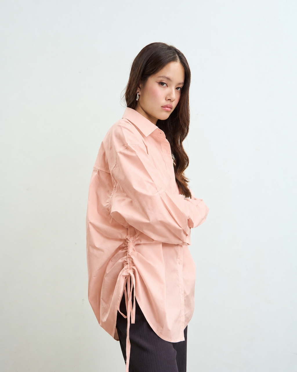 woman wearing a old rose long-sleeve button-up shirt with side drawstrings and dark pants, standing against a plain light gray background.